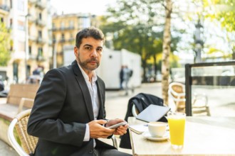 Elegant adult caucasian businessman using phone while drinking coffee in an outdoors cafeteria in