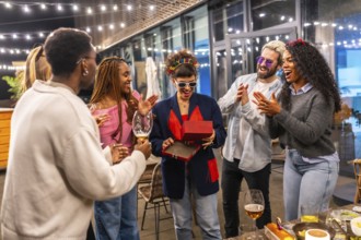 Young multi-ethnic friends giving birthday gift to a woman at night party in the terrace of a bar