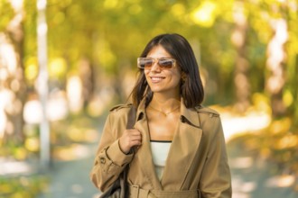 A woman wearing sunglasses and a brown coat is walking down a path. She is smiling and she is