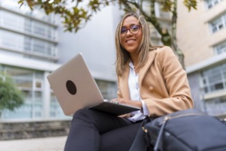 Happy latin businesswoman working with laptop sitting in the city next to a financial building