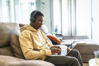 Young man sitting comfortably on a sofa, using a laptop and headphones, focused on studying and