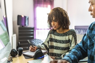 Two programmers working from home, writing notes on notepad while looking at computer screen