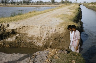 Two men stand on a broken country road next to a canal in rural Pakistan in 1962