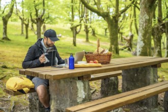 A young Caucasian father teleworking sitting at a picnic table with computer in the woods. Summer