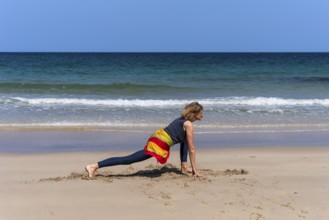 A woman doing yoga on a sandy beach by the sea. Ashwa Sanchalanasana or Sprinter, variant. Treen