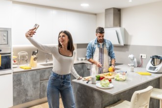 Happy couple preparing healthy meal in their kitchen, woman taking selfie while man is cutting