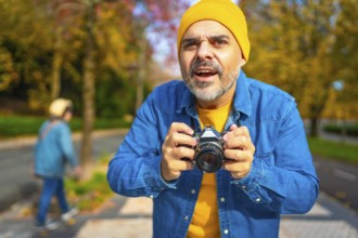 Surprised mature caucasian man holding digital camera about to photographing in the city in autumn