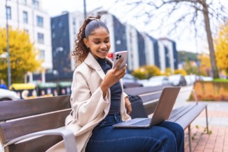Happy african american woman using smartphone and laptop sitting on a city bench in autumn