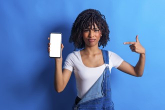 Young woman showing a smartphone with blank white screen and pointing at it with index finger,