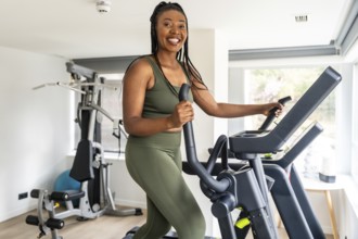 Happy african american woman exercising on elliptical cross trainer in bright home fitness room