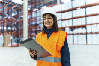 Warehouse worker wearing safety vest and helmet smiling and holding clipboard in logistics center