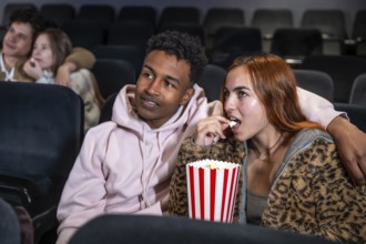 Young multi ethnic couple enjoying a movie and sharing popcorn in a cinema, surrounded by other