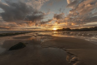 A beach with a rock and a sunset, the sky is reflected on the sand of a beach. The sky is cloudy