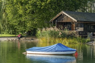 Wooden hut with common reed (Phragmites australis), covered boat, two woman on a park bench, shore