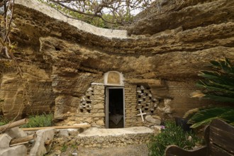 Exterior view of a cave carved into the rock with a stone wall and vegetation around the entrance,