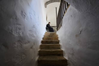 A monk sits on a bench at the end of a stone staircase in a narrow corridor of the monastery,