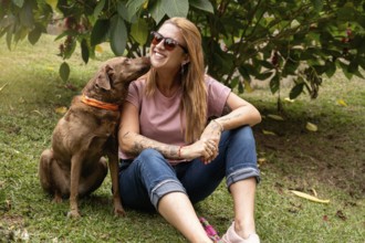 The happy woman with sunglasses and her dog sitting in the park, her pet gives her a kiss on the