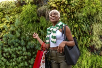 African woman looking at camera while carrying shopping bags next to a garden