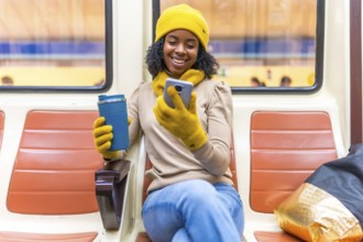 Smiling black woman wearing yellow winter clothes using phone and drinking coffee while traveling