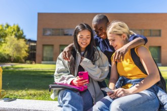 Happy multi-ethnic students using mobile and sitting outside the university campus in a sunny day
