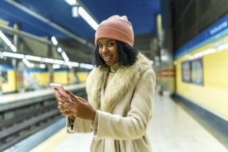 Stylish young woman using mobile phone while waiting for subway train, enjoying her commute in