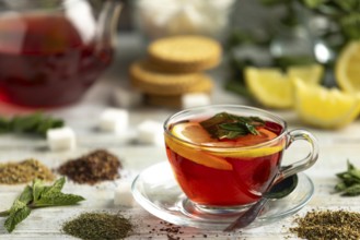A cup of red tea with lemon and mint surrounded by sugar cubes, teas and biscuits