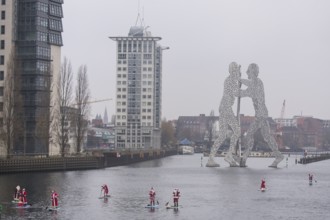 Water sports enthusiasts dressed as Santas ride SUPs on the Spree in front of the Molecule Man in