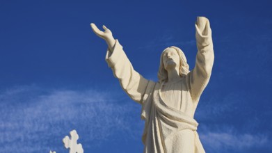 A stone statue of Jesus with outstretched arms against a blue sky, Agios Nikolaos church, cemetery,
