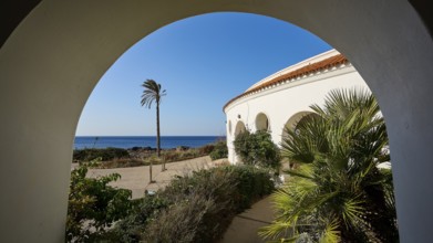 View through an arch of a circular building to the sea, surrounded by green plants and bushes,