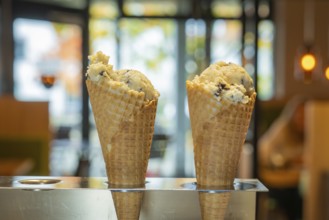 Two ice cream cones in the foreground in front of a warm café ambience with glass windows and soft