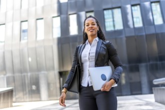 Professional young latin businesswoman standing carrying laptop in the city next to a modern