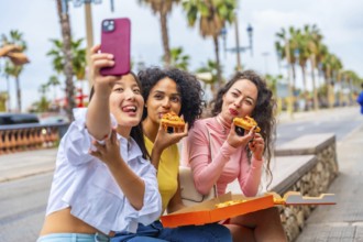 Multiracial female friends eating pizza and taking a selfie sitting in the street