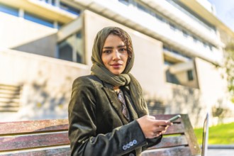 Muslim businesswoman using smartphone sitting on a park bench next to office building