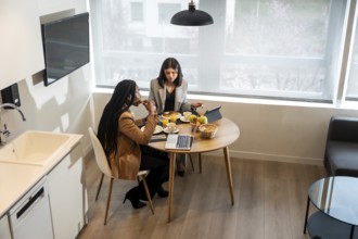 Two businesswomen are having breakfast in their hotel room while working on a tablet and laptop