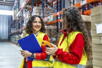 Two young women talking while taking inventory in a distribution warehouse using clipboard and