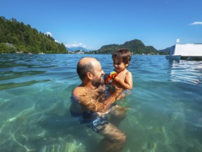 Father playing with his son in the clear water of lake bled, with mountains and forest in the