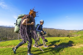 Two female climbers are walking across a mountain meadow carrying climbing rope and gear on a sunny