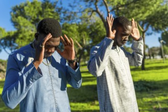 Two young senegalese men wearing traditional dashiki clothing are praying outdoors in a park,