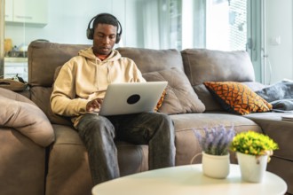 Young man concentrating on his laptop while wearing headphones, comfortably seated on a sofa in his