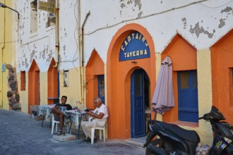 People sitting in front of a colourful facade of a taverna and enjoying their time on the street,