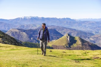 Male hiker walking on grassy mountain slope with trekking poles, enjoying scenic view of mount