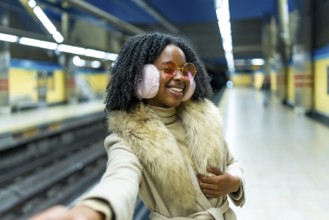 Young black woman wearing earmuffs and fur, taking a selfie with her smartphone in a subway station