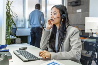 Young asian manager working diligently at a computer, wearing a headset in a sleek, modern