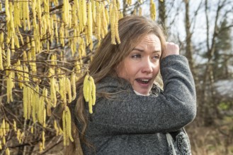 Young Caucasian woman with allergy problem stands by flowering hazel bush and sneezes in the crook