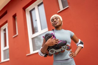 Confident and proud african woman carrying inline skates on shoulders in the city next to house