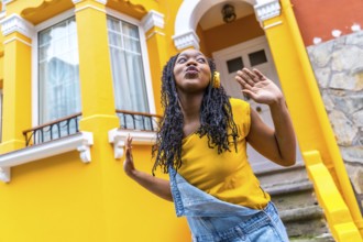 Cute african woman dancing listening to music with headphones next to a yellow house outdoors