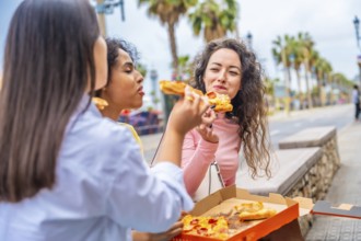 Three female diverse tourists chatting and sharing pizza sitting in the street