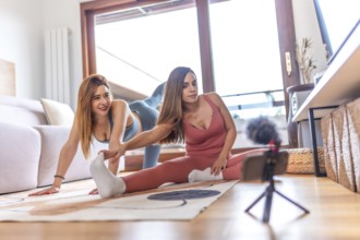 Two young women recording an online yoga class at home, performing stretching exercises in the