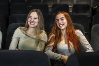 Two happy young women are sitting in comfortable cinema seats, enjoying a movie and smiling