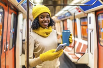 Smiling woman wearing yellow winter clothes using smartphone and drinking coffee inside subway